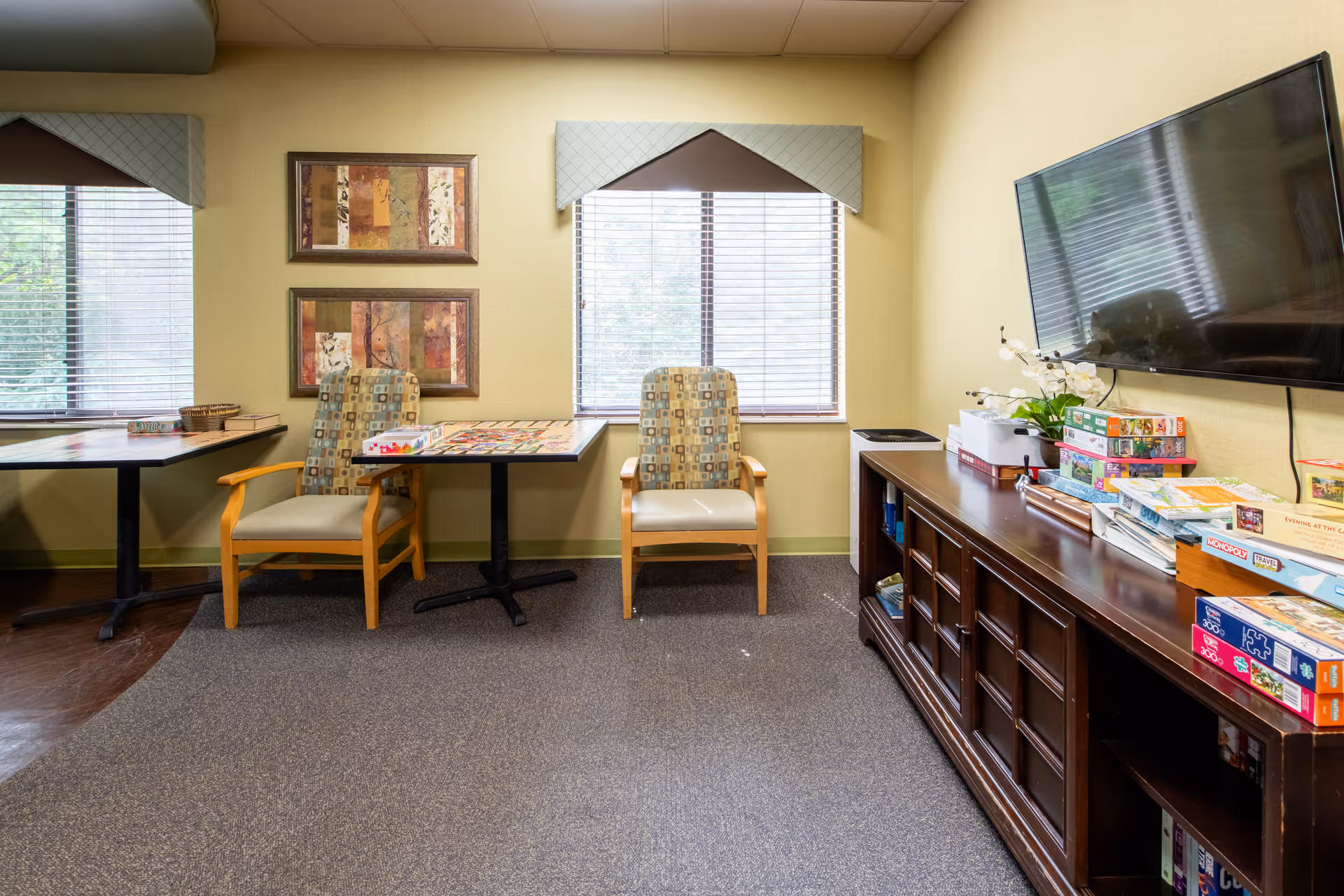 A cozy common area with two patterned armchairs and a table set up for board games. The room has two windows with blinds and valances, two framed abstract paintings on the wall, a large flat-screen TV mounted on the wall, and a dark wooden cabinet filled with board games and books.