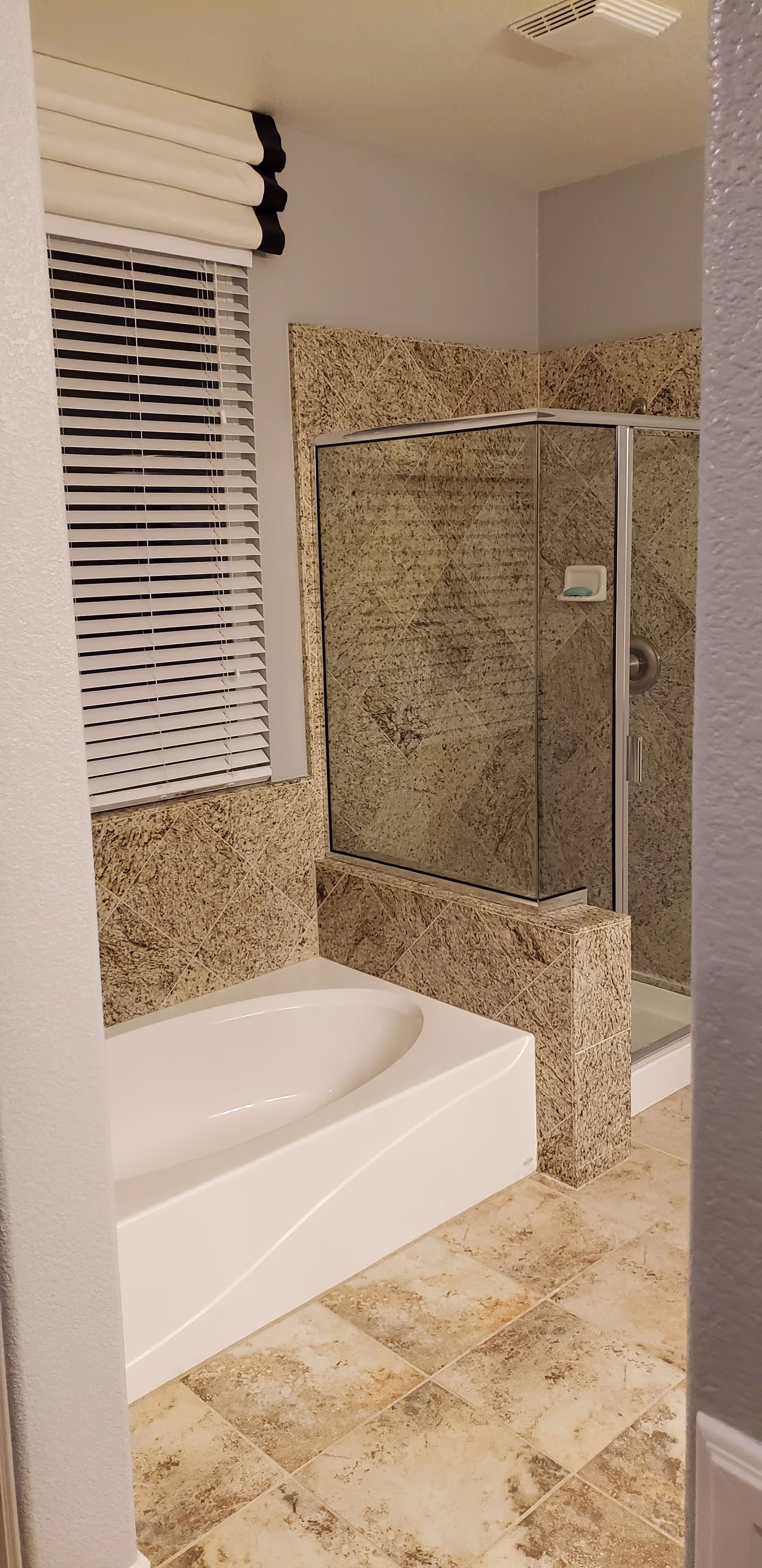 Bathroom with a white bathtub and a glass-enclosed shower. The walls and floor around the bathtub and shower are covered with beige and brown patterned tiles. There is a window with white blinds and a white and black valance above the bathtub.