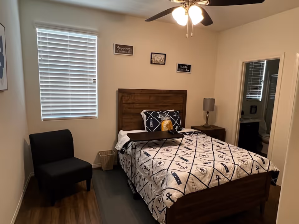 A bedroom with a wooden bed frame and nautical-themed bedding. There is a black chair to the left, a small nightstand with a lamp to the right, and a ceiling fan with lights above. A window with closed blinds is on the left wall, and three small decorative signs are hung above the bed. An open door on the right leads to a bathroom.