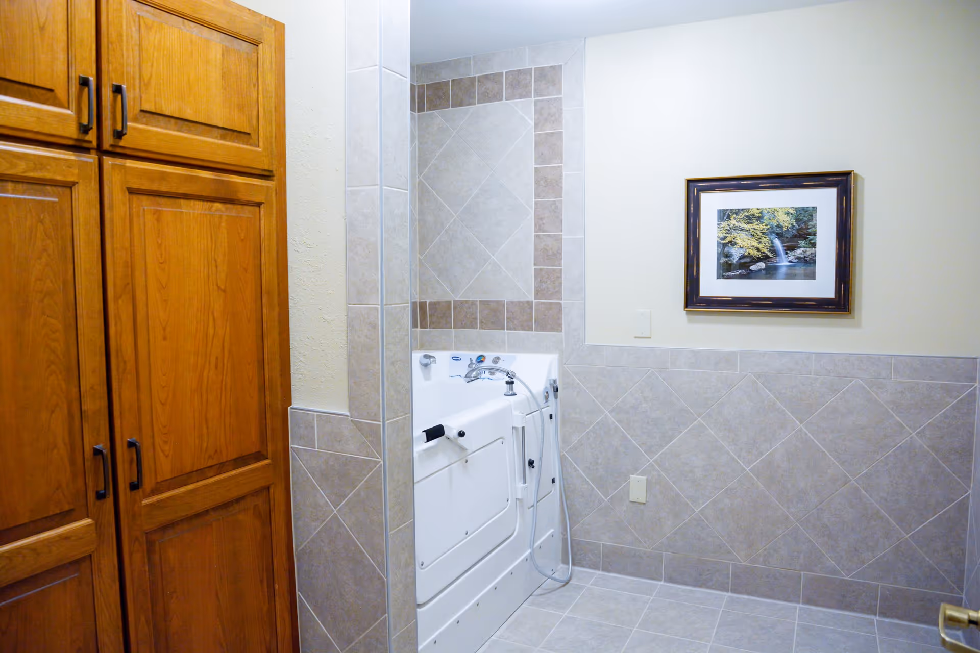A bathroom with beige tiled walls and floor, featuring a white walk-in bathtub with a faucet and handheld shower. There is a wooden cabinet on the left side and a framed picture of a waterfall on the wall above the bathtub.