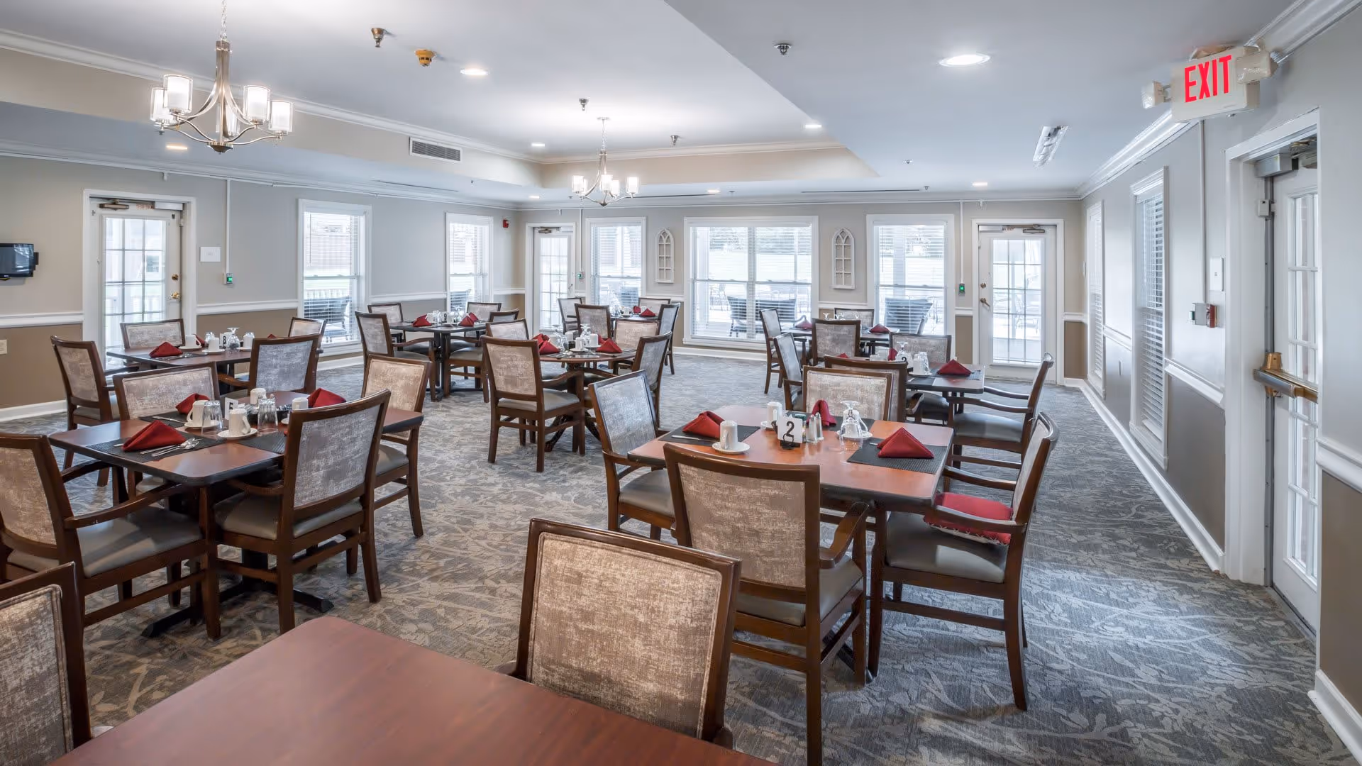 A spacious dining room with multiple wooden tables and chairs arranged neatly. Each table is set with red folded napkins, cups, and small condiment containers. The room has large windows allowing natural light to fill the space, light gray walls with white trim, and two chandeliers hanging from the ceiling. There are exit doors visible on the right side and at the back of the room.