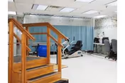 Interior view of a rehabilitation or physical therapy room featuring wooden stairs with handrails for practice, exercise equipment including a recumbent bike, and a desk with chairs and a computer in the background.