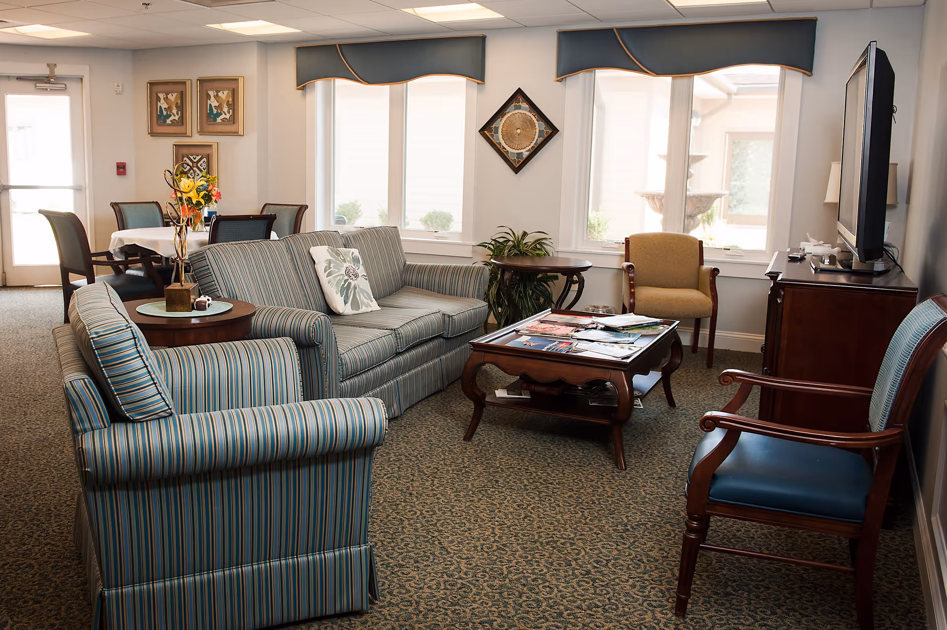 A cozy senior living common area with striped upholstered sofas and chairs arranged around a wooden coffee table holding magazines. There is a TV on a wooden stand to the right, a small round side table with a plant, and a dining table with chairs in the background near a door. The room has large windows with blue valances and framed artwork on the walls.