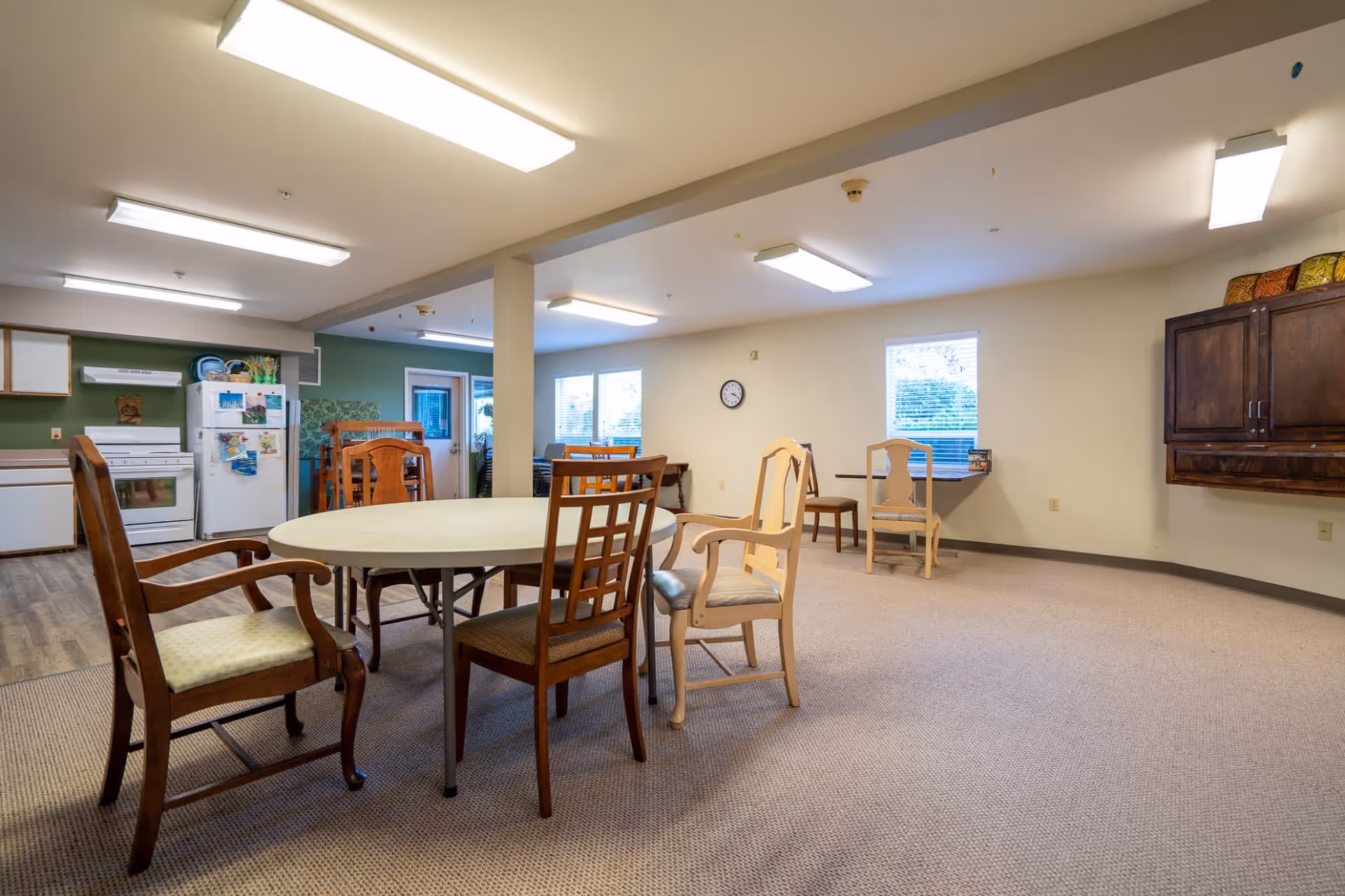 A spacious common area in a retirement facility featuring a round table with various wooden chairs around it. In the background, there is a kitchen area with a white refrigerator and stove, green walls, and a door leading outside. The room has beige carpeting, fluorescent ceiling lights, and windows letting in natural light.
