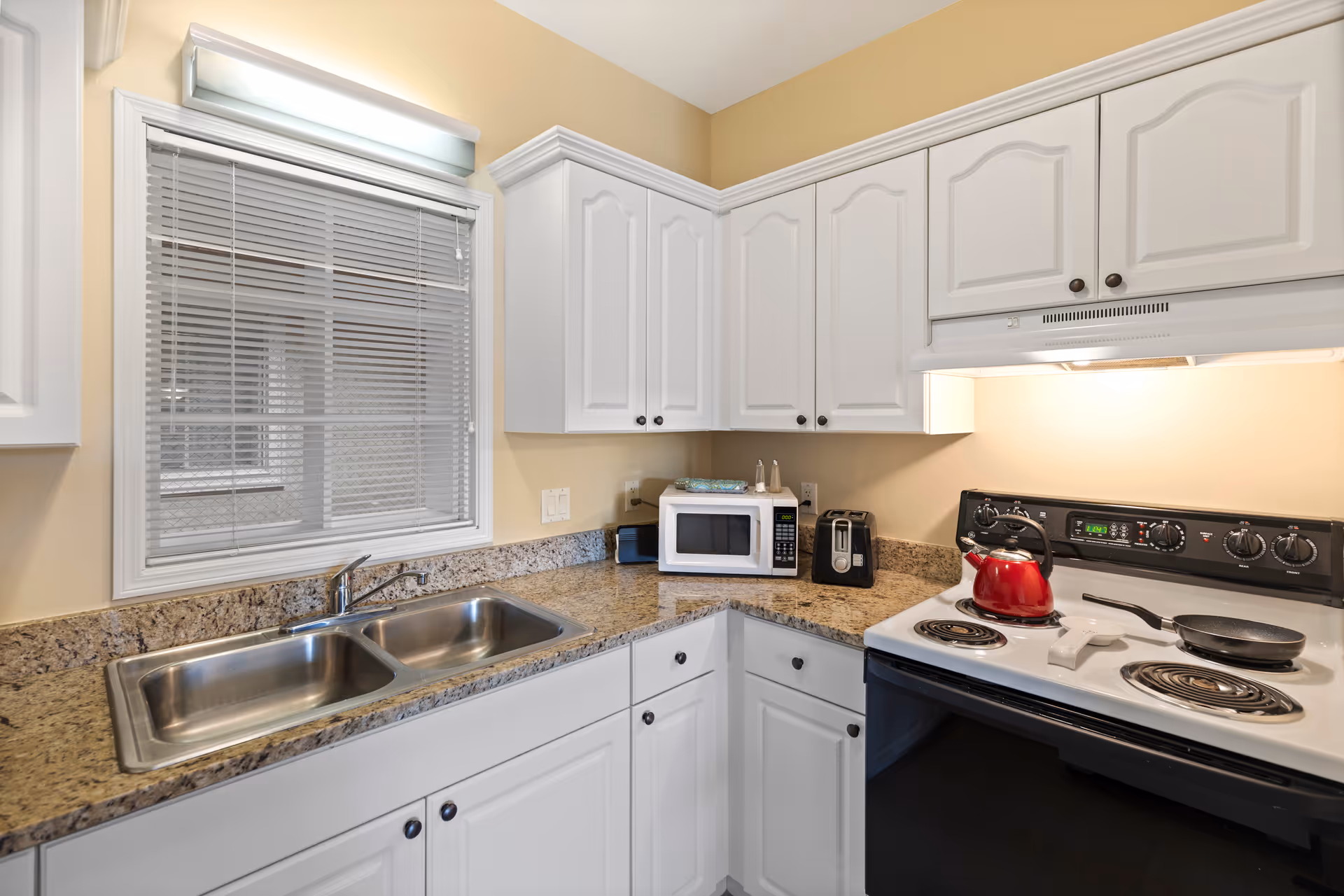 Bright kitchen with white cabinets, granite countertops, a double stainless sink, microwave, and electric stove with a red kettle.