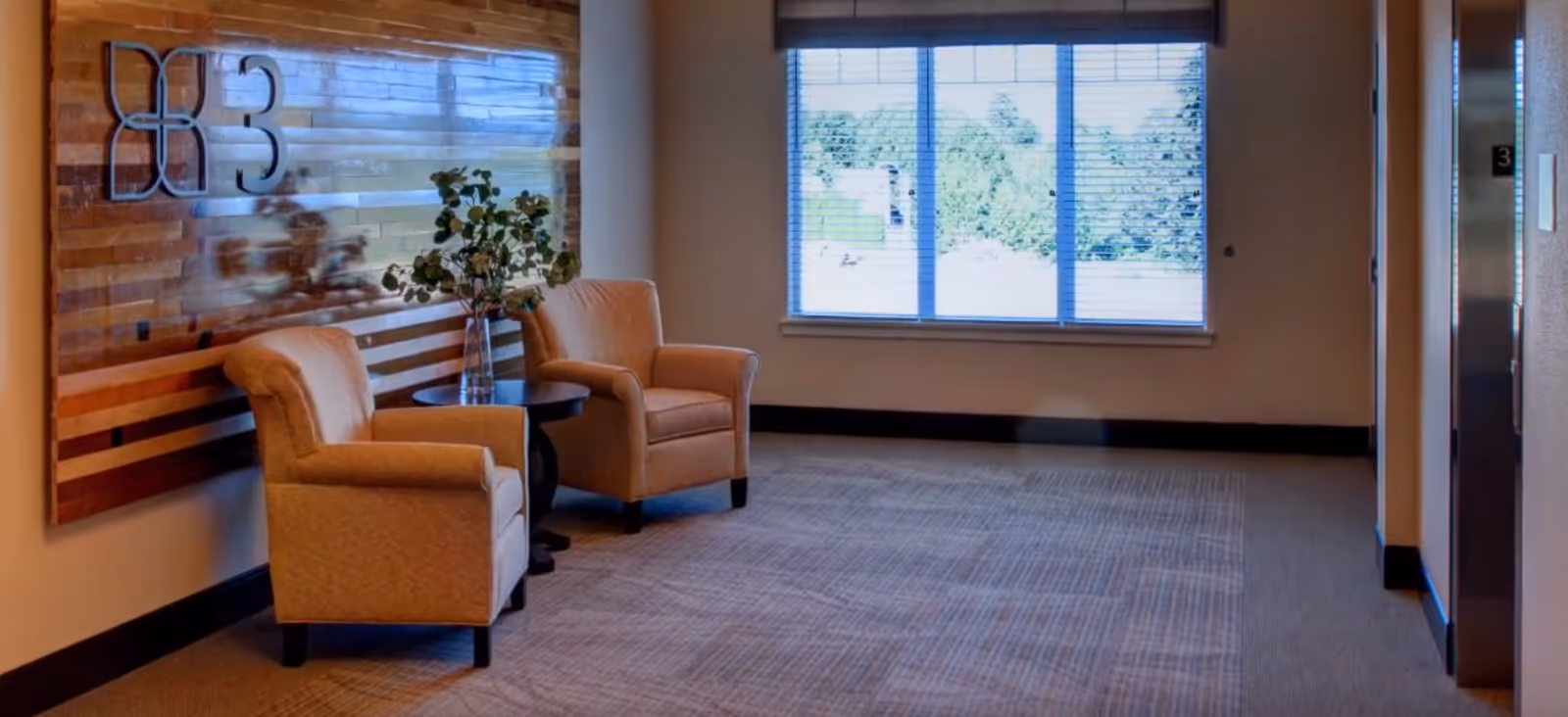 A quiet interior common area with two upholstered chairs and a small table in front of a window and wooden wall art near an elevator.