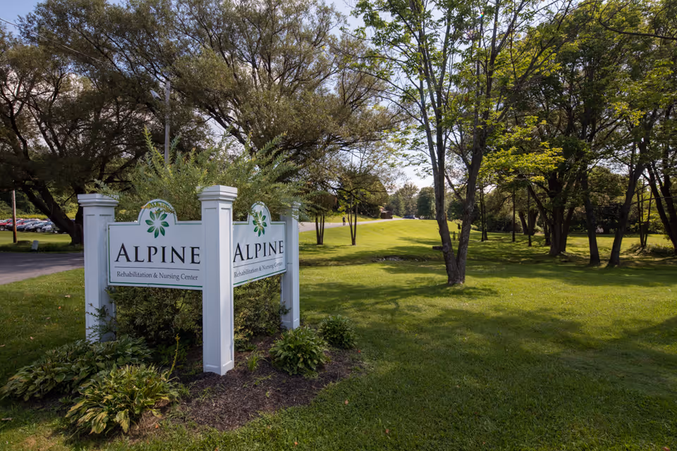Outdoor view of a green grassy area with trees and a white sign that reads 'Alpine Rehabilitation & Nursing Center' surrounded by bushes and plants.