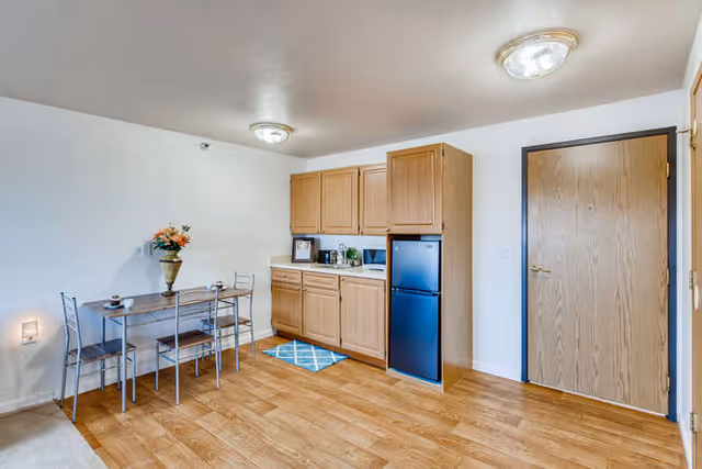 Small studio kitchenette with oak cabinets, a compact refrigerator and microwave, and a small dining table with chairs against a white wall.