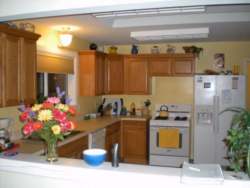 A kitchen with wooden cabinets, a white stove with a yellow towel hanging on the handle, a white refrigerator with magnets and notes, a dishwasher, and a countertop with a vase of colorful flowers, a blue bowl, and various kitchen items. There is a window with a closed blind and overhead fluorescent lighting.