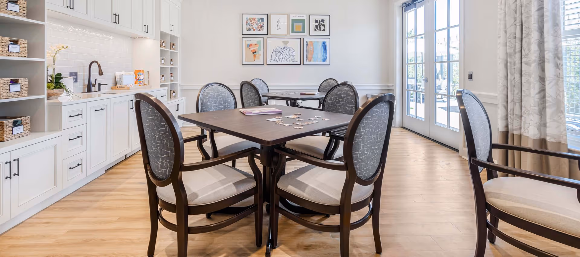 A bright and clean room with wooden flooring featuring two square tables surrounded by cushioned chairs with wooden frames. On the tables, there are puzzle pieces and books. The room has white cabinetry with baskets and a sink on the left side. The back wall is decorated with framed abstract artwork, and large glass doors on the right let in natural light.