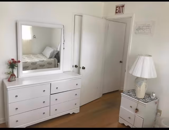 A bright bedroom with white walls and wooden floor. There is a white dresser with six drawers and a large mirror on top, reflecting a bed with a patterned bedspread and pillows. A small vase with red flowers is placed on the dresser. To the right, there is a white nightstand with two drawers, a decorative lamp with a white shade, and a small clock. Two white doors are visible, one slightly open, and an exit sign is mounted above one door.