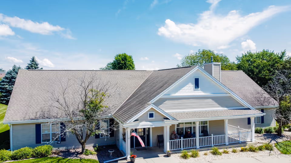 Front exterior of a single-story senior living residence with a covered porch, American flag, and landscaped yard under a blue sky.
