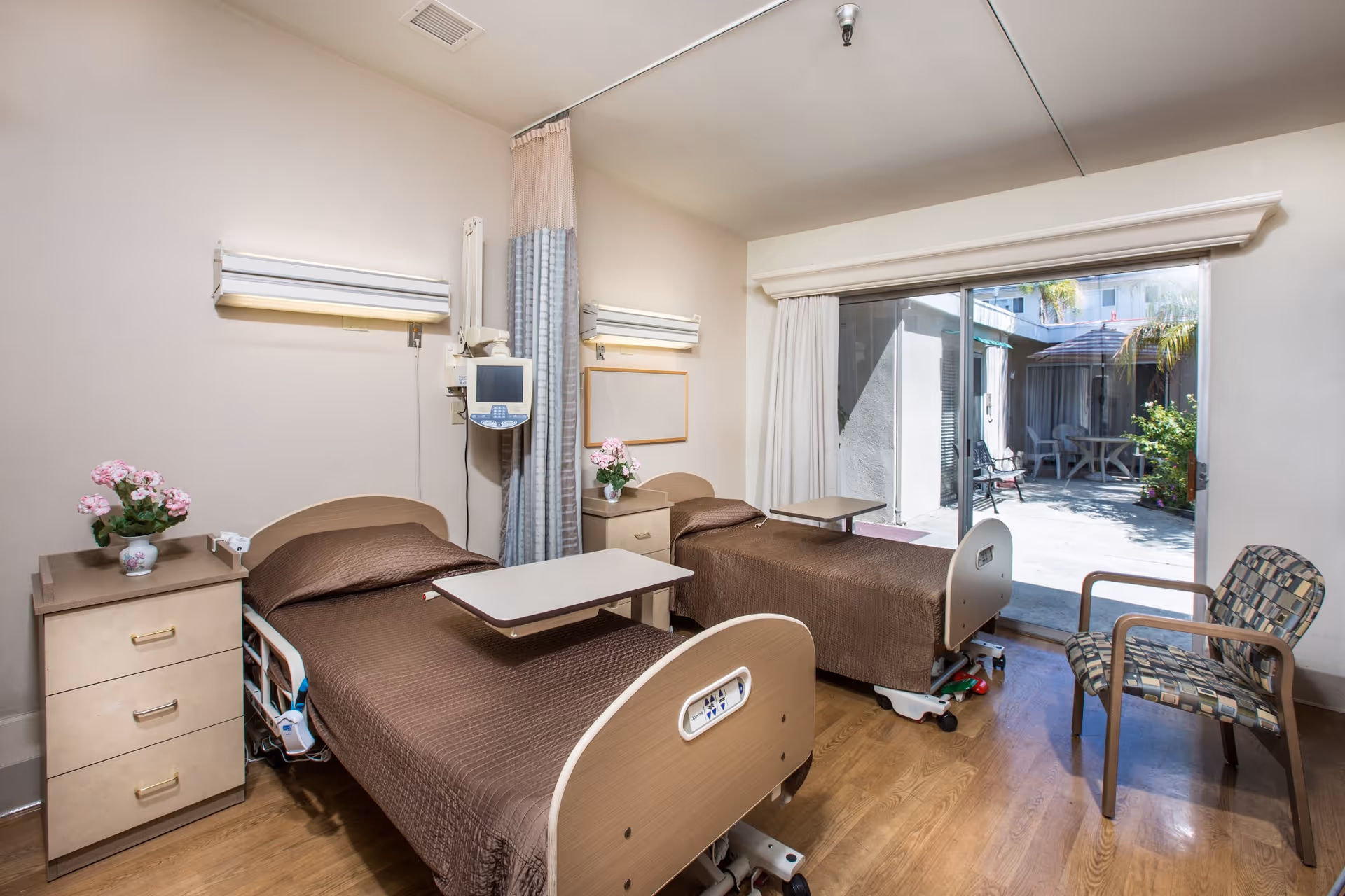 A room with two hospital-style beds covered with brown bedspreads, each with an overbed table. There are two bedside cabinets with pink flowers in vases on top. A medical monitor is mounted on the wall between the beds, and a privacy curtain hangs between them. A sliding glass door leads to an outdoor patio area with chairs and a table under an umbrella. A patterned armchair is positioned near the door.