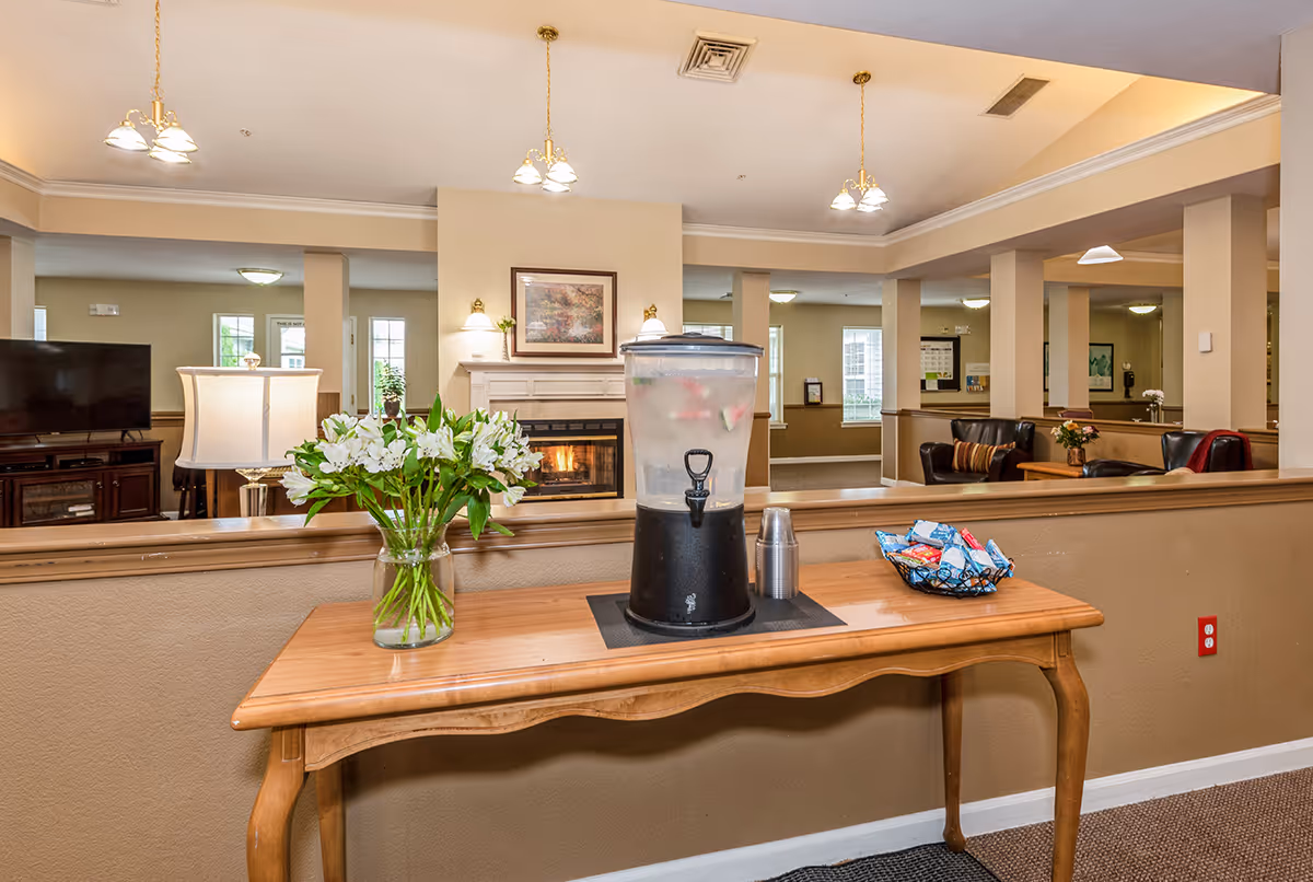 Communal lounge area with a wooden table holding a beverage dispenser, vase of flowers, and snacks, with seating and a fireplace visible in the background.