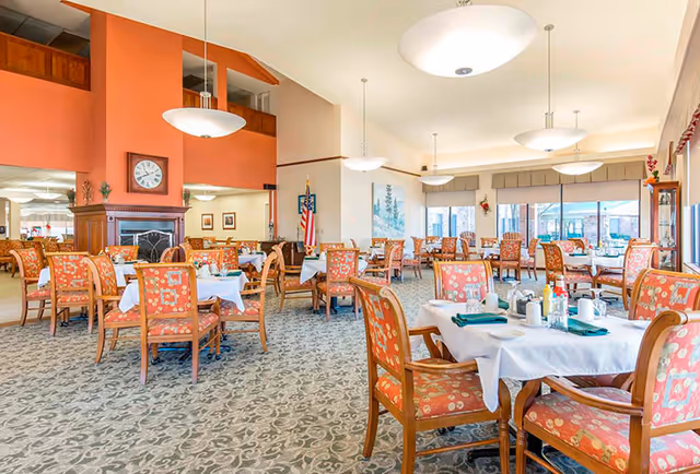 A spacious dining room with multiple tables covered in white tablecloths, each set with plates, cups, and green napkins. The chairs have wooden frames with orange and floral patterned upholstery. The room features large windows allowing natural light, a fireplace with a clock above it, and several hanging ceiling lights. An American flag is displayed near the fireplace.