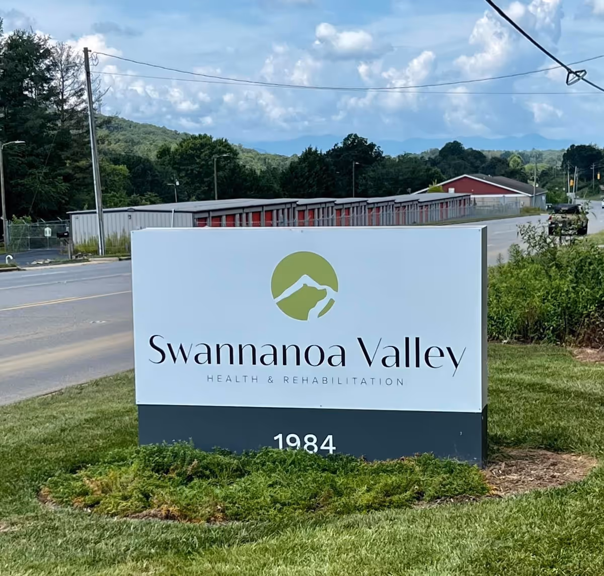Outdoor view of a sign for Swannanoa Valley Health & Rehabilitation located on a grassy area beside a road, with trees, storage units, and mountains visible in the background under a partly cloudy sky.