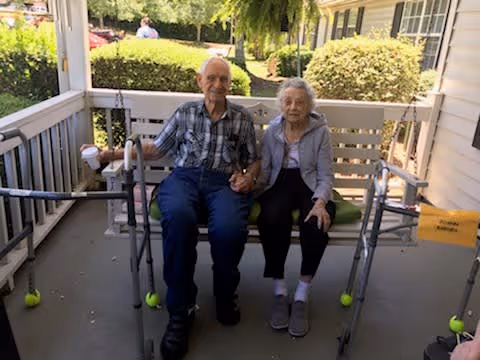 An elderly man and woman sitting together on a white porch swing outside a building. Both have walkers with tennis balls on the legs positioned in front of them. The porch is surrounded by white railings and greenery is visible in the background.
