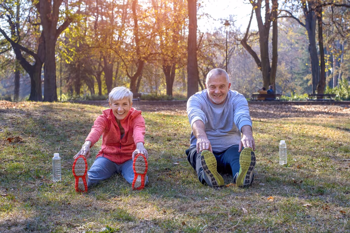 An elderly woman and man sitting on grass in a park, stretching forward to touch their toes with water bottles placed beside them, surrounded by trees with autumn foliage.
