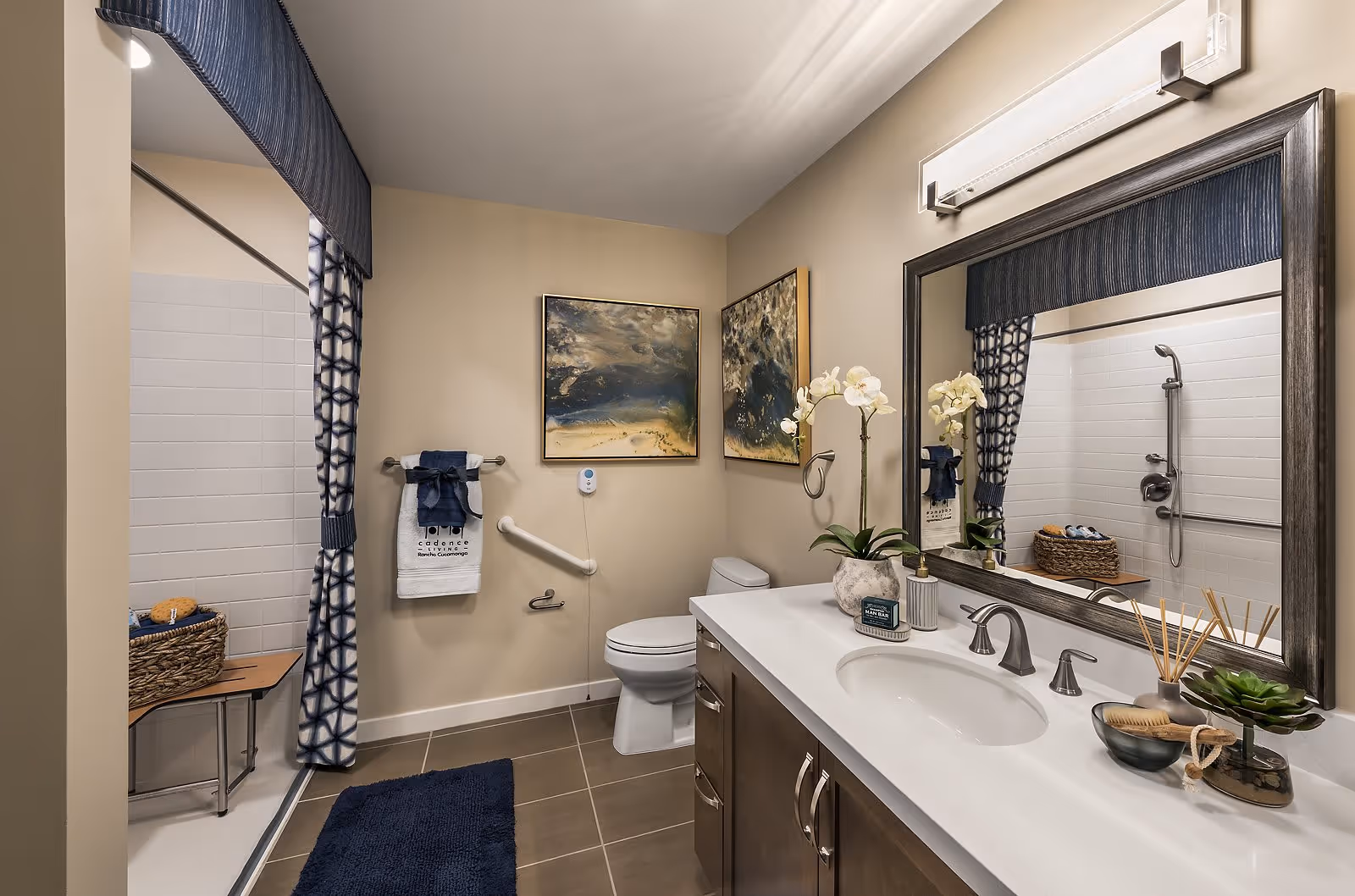 A modern bathroom with a white toilet, a large mirror above a white countertop with a sink, and a wooden cabinet below. The shower area has a blue and white patterned curtain and a foldable wooden bench. There are two abstract paintings on the beige wall, a blue bath mat on the floor, and decorative plants and toiletries on the countertop. A towel with the text 'Cadence at Rancho Cucamonga' hangs on a towel rack.