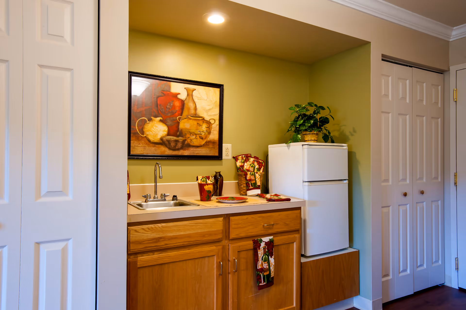 Small kitchenette area with a white mini refrigerator, wooden cabinets, a sink with a faucet, and a countertop decorated with a plant, a framed painting of pottery on the wall, and some kitchen towels and utensils.