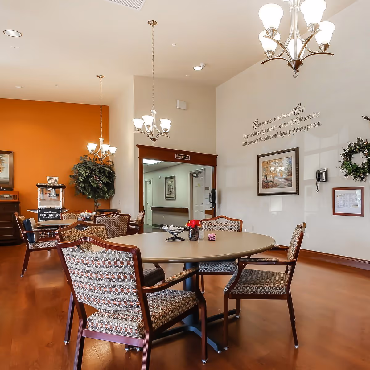 A well-lit dining area in an assisted living facility featuring a round table with six patterned chairs. The room has warm wooden flooring, two chandeliers, a popcorn machine, a large plant, and framed artwork on the walls. There is a hallway visible through an open doorway, and a wall decal with an inspirational message about honoring God and providing high-quality senior lifestyle services.