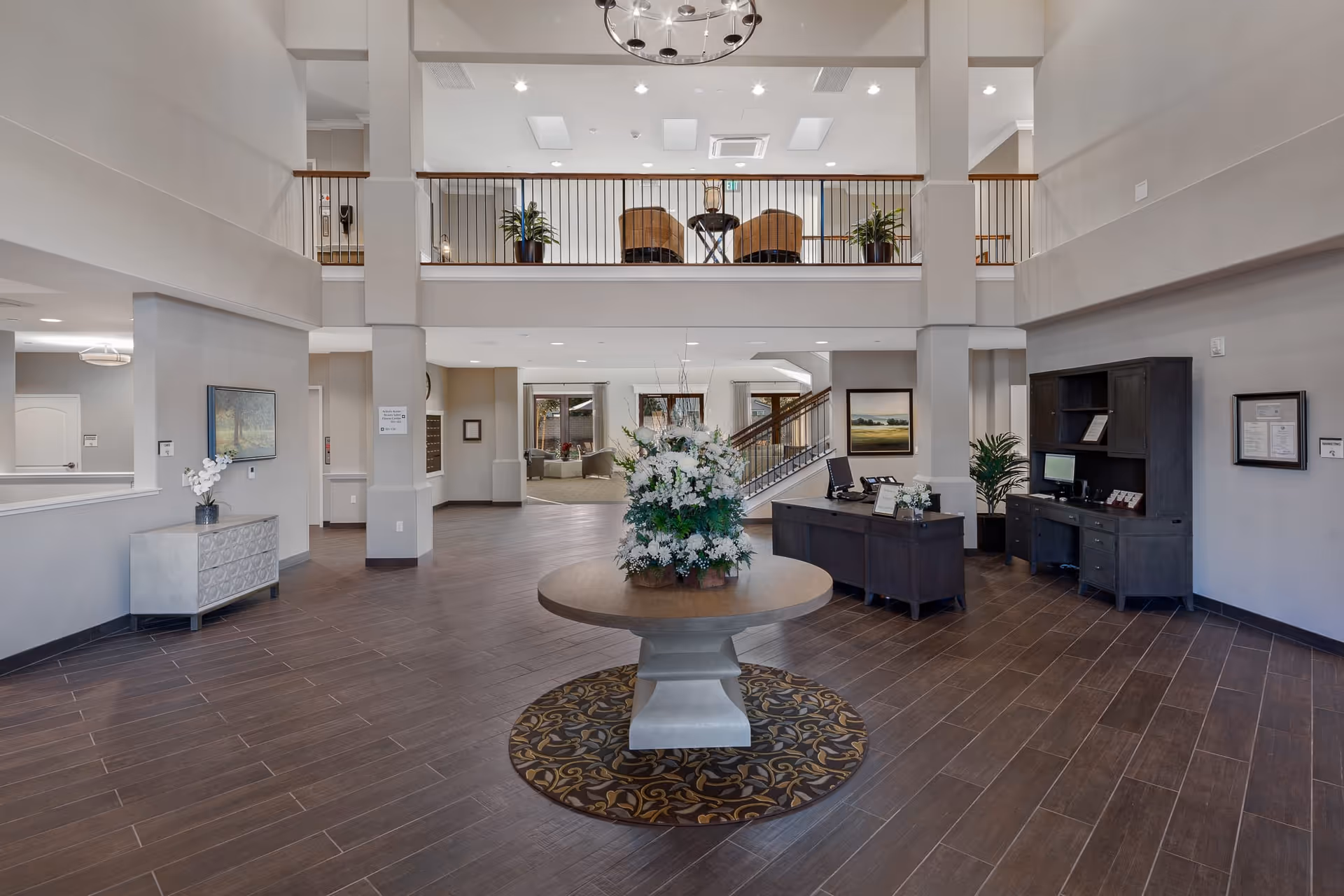 Spacious and well-lit senior living facility lobby with a round table featuring a large floral arrangement in the center. The lobby has dark wood flooring, a reception desk with computer and phone on the right, and a seating area visible in the background. There is a second-floor balcony with chairs and plants overlooking the lobby.