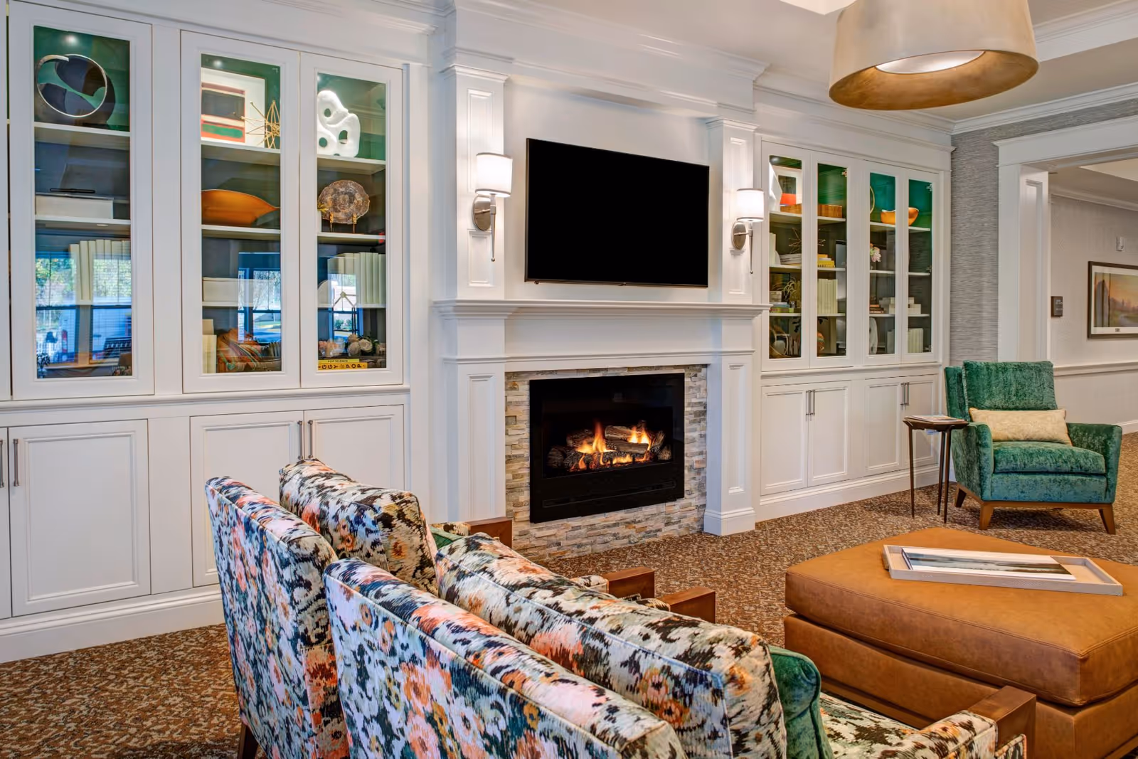 Bright community living room with floral-upholstered chairs facing a fireplace and wall-mounted TV, flanked by white built-in glass cabinets and additional seating.