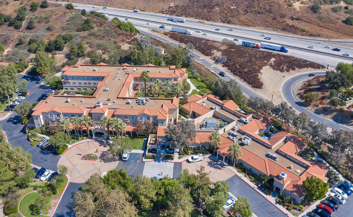 Aerial view of the Bayshire San Dimas senior living complex with red tile roofs, landscaping, parking areas, and a nearby freeway.