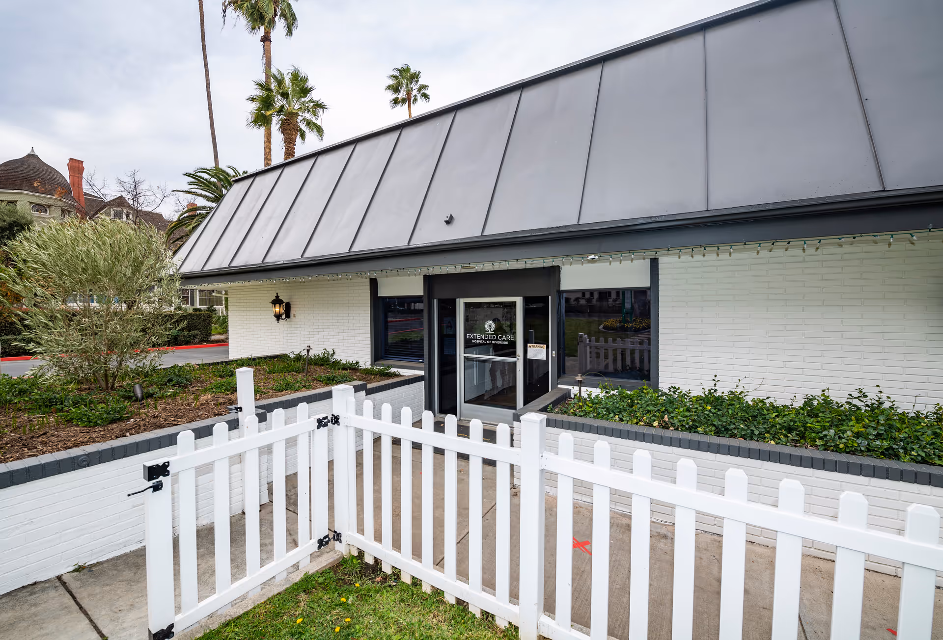 Front entrance of Extended Care Hospital of Riverside with a white picket fence, landscaped beds, and a glass double door.