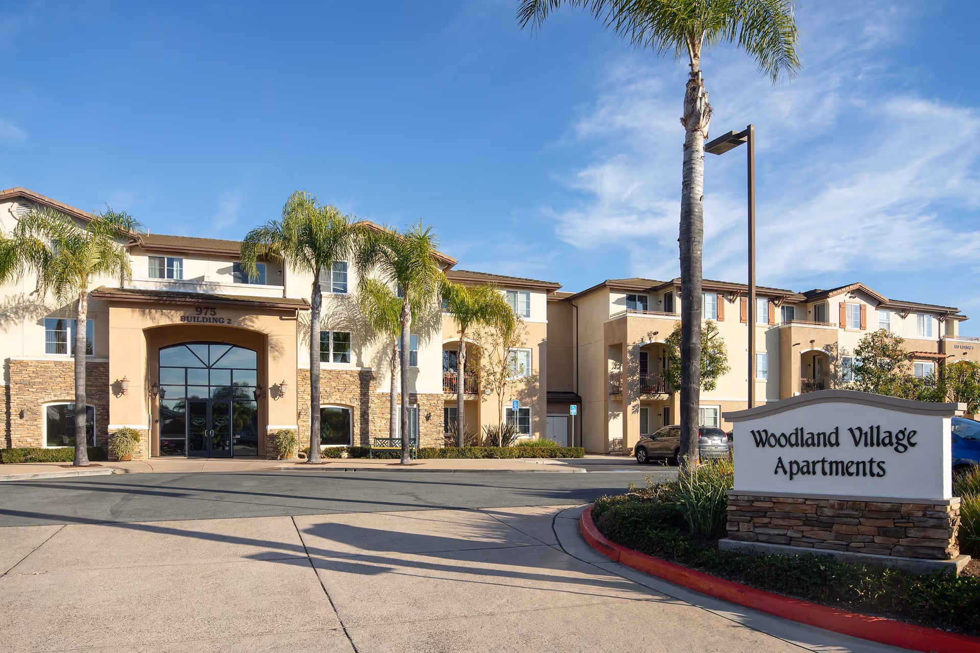 Exterior view of Woodland Village Apartments, a multi-story residential building with beige and stone facade, palm trees, and a clear blue sky. A sign in front reads 'Woodland Village Apartments'.