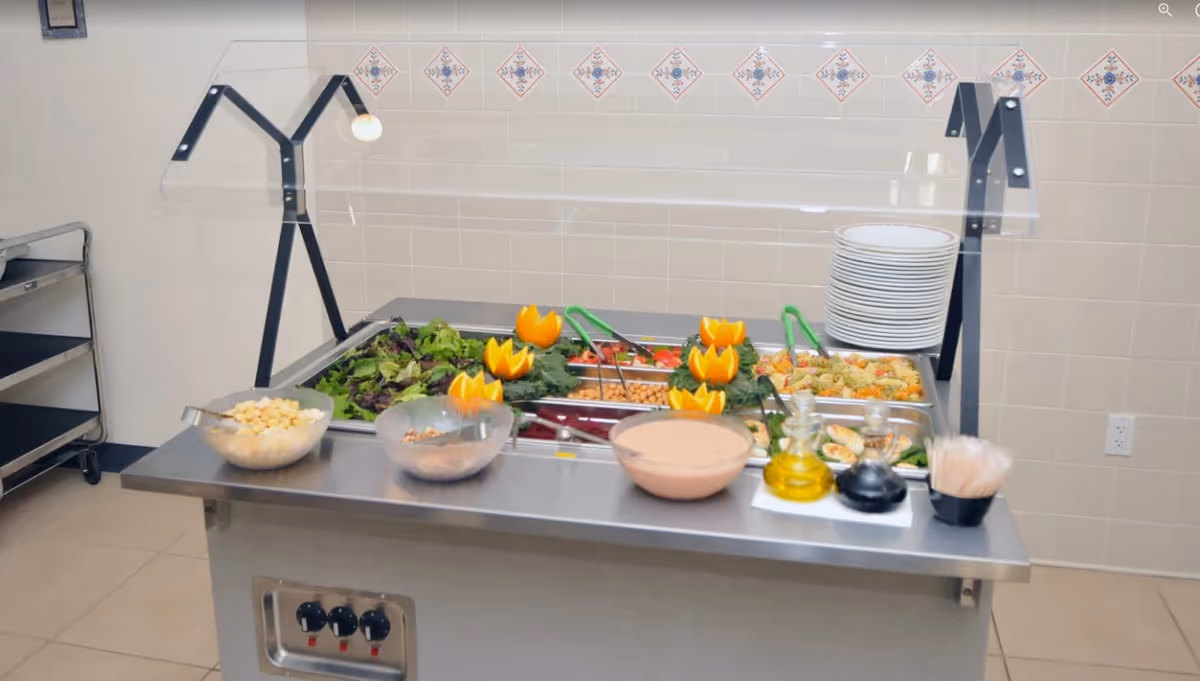 A stainless-steel salad buffet station with bowls of salad, dressings and stacked plates against a tiled wall.