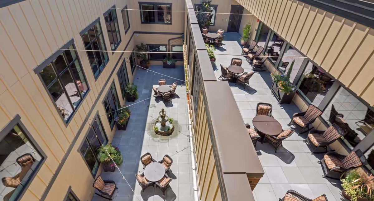 Aerial view of an outdoor courtyard area in a senior living facility with multiple round tables and cushioned chairs arranged on a tiled floor. The courtyard is surrounded by two-story building walls with windows and potted plants along the edges. String lights are hung across the courtyard, and a decorative fountain is visible in the center.