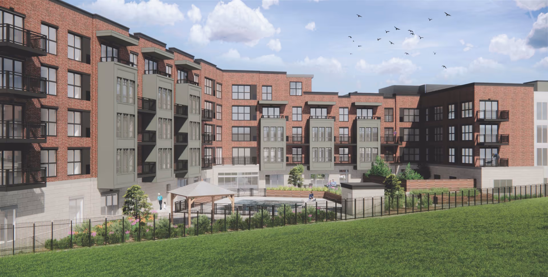 Exterior view of a multi-story senior living facility with red brick and gray accents, featuring balconies and large windows. The foreground shows a fenced garden area with a gazebo, greenery, and a grassy lawn under a partly cloudy sky with birds flying.