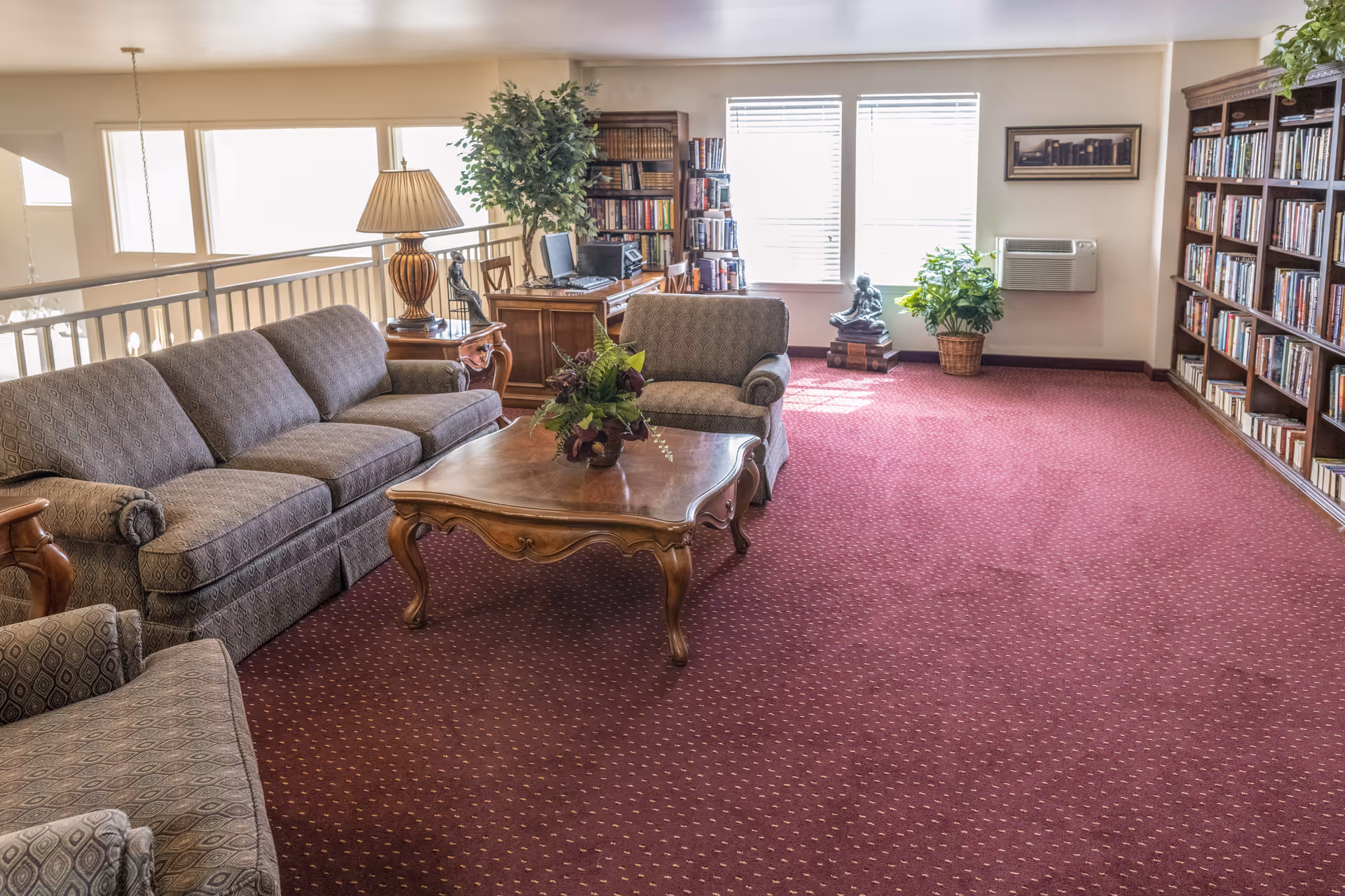 A cozy living room area with patterned upholstered sofas and armchairs surrounding a wooden coffee table with a floral arrangement. The room has red carpet flooring, large windows letting in natural light, bookshelves filled with books, a wooden desk with a computer, a table lamp, and potted plants near the windows.