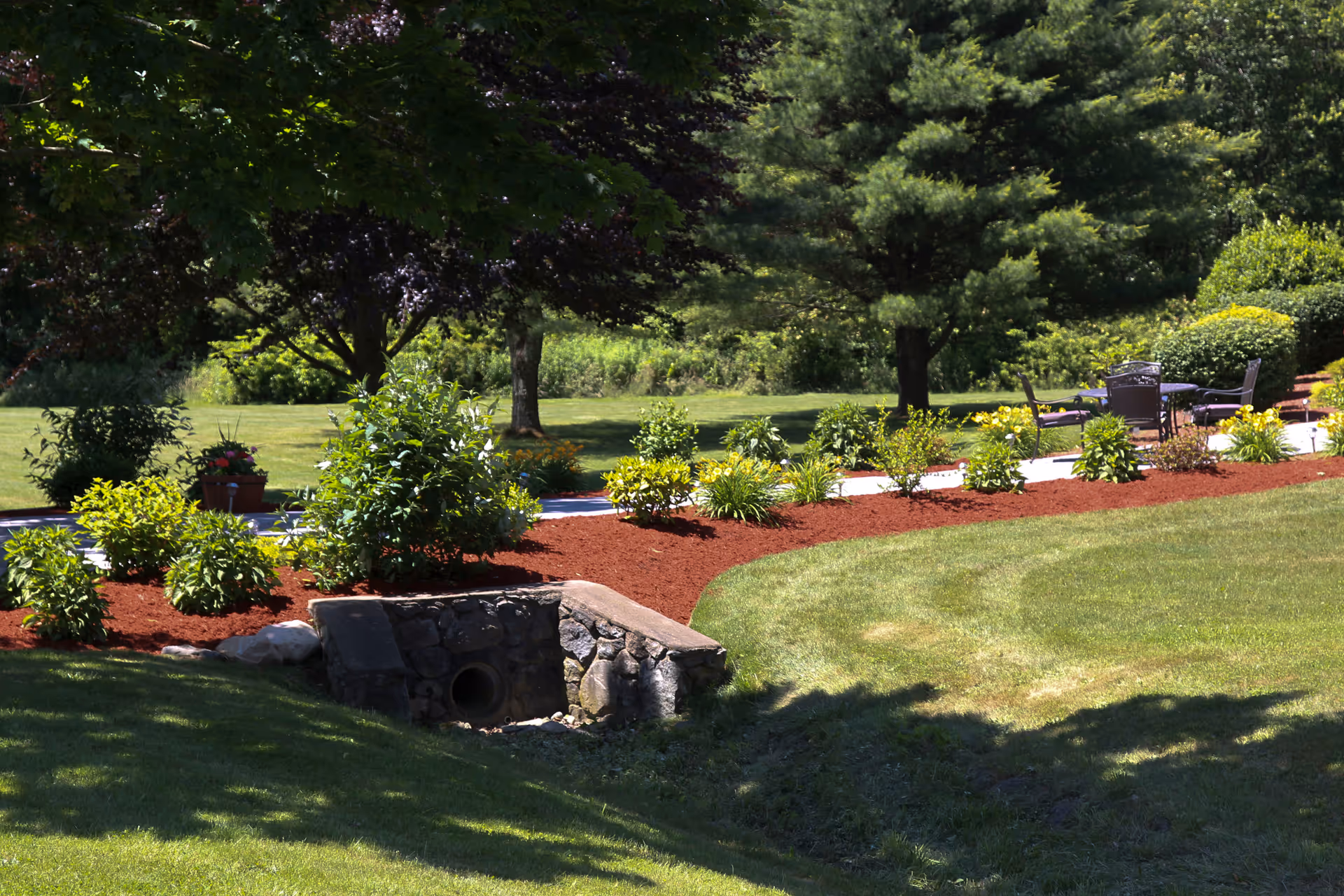 A landscaped outdoor garden area with green grass, various shrubs and plants bordered by red mulch, a stone drainage pipe, and several trees providing shade. There is a paved walkway with outdoor chairs and a table in the background.