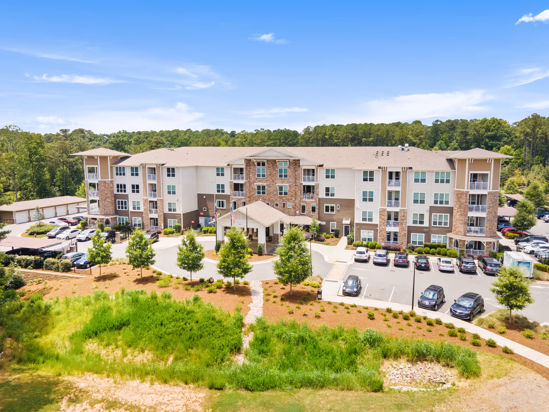Aerial view of Verena at Bedford Falls, a modern four-story senior living facility surrounded by greenery and trees. The building features a combination of stone and beige siding with multiple balconies. There is a circular driveway with a covered entrance, several parked cars, and landscaped grounds with small trees and shrubs under a blue sky with some clouds.