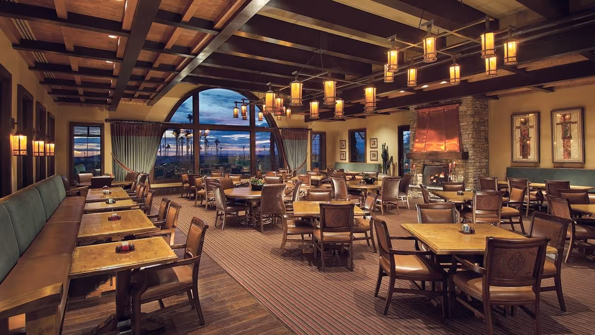 Warm, spacious dining room with wooden tables and chairs, a stone fireplace, hanging lights, and large arched windows showing an evening view.