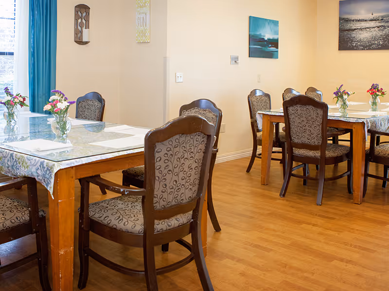 A dining room with wooden tables covered with floral tablecloths and glass tops. Each table has several wooden chairs with patterned upholstery. Small vases with colorful flowers are placed on the tables. The room has light-colored walls with framed artwork and a window with blue curtains letting in natural light.