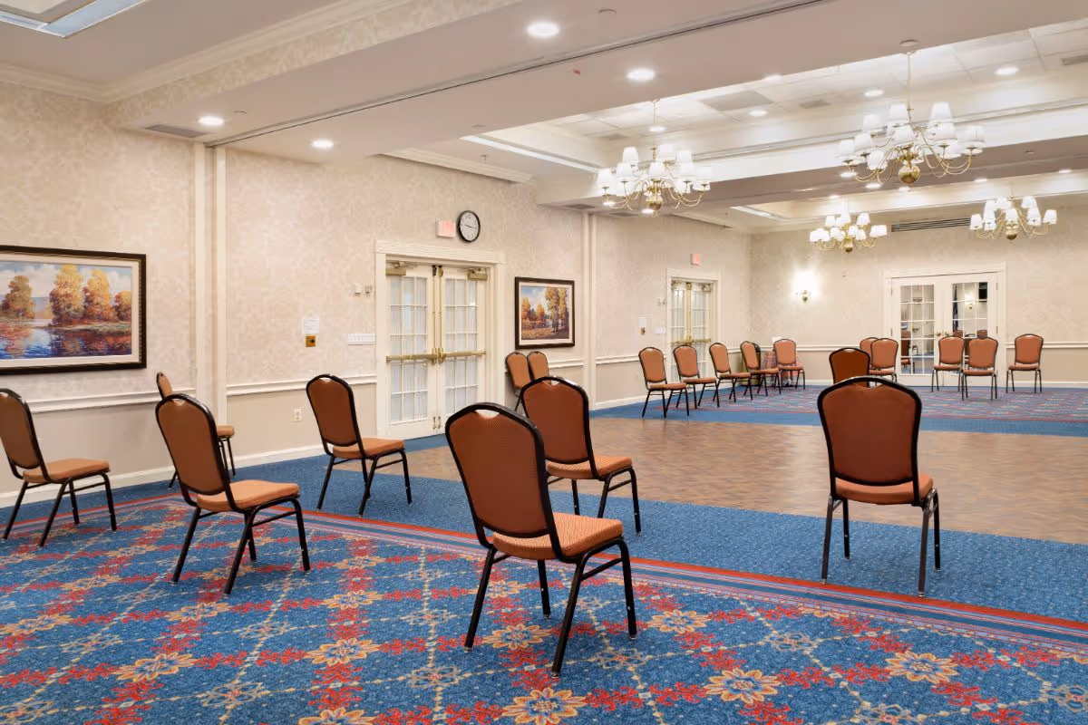 Spacious multipurpose event room with rows of spaced chairs on patterned carpet and a wooden floor area under chandeliers.