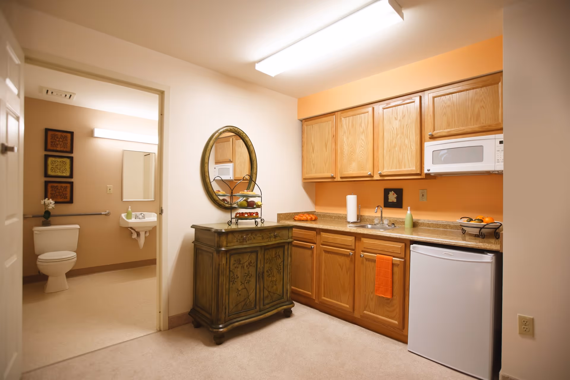 Interior view of a kitchenette area with wooden cabinets, a small white refrigerator, a microwave, a sink, and a countertop with a paper towel holder and fruit bowls. To the left, there is an open doorway leading to a bathroom with a toilet, sink, mirror, and wall decorations.