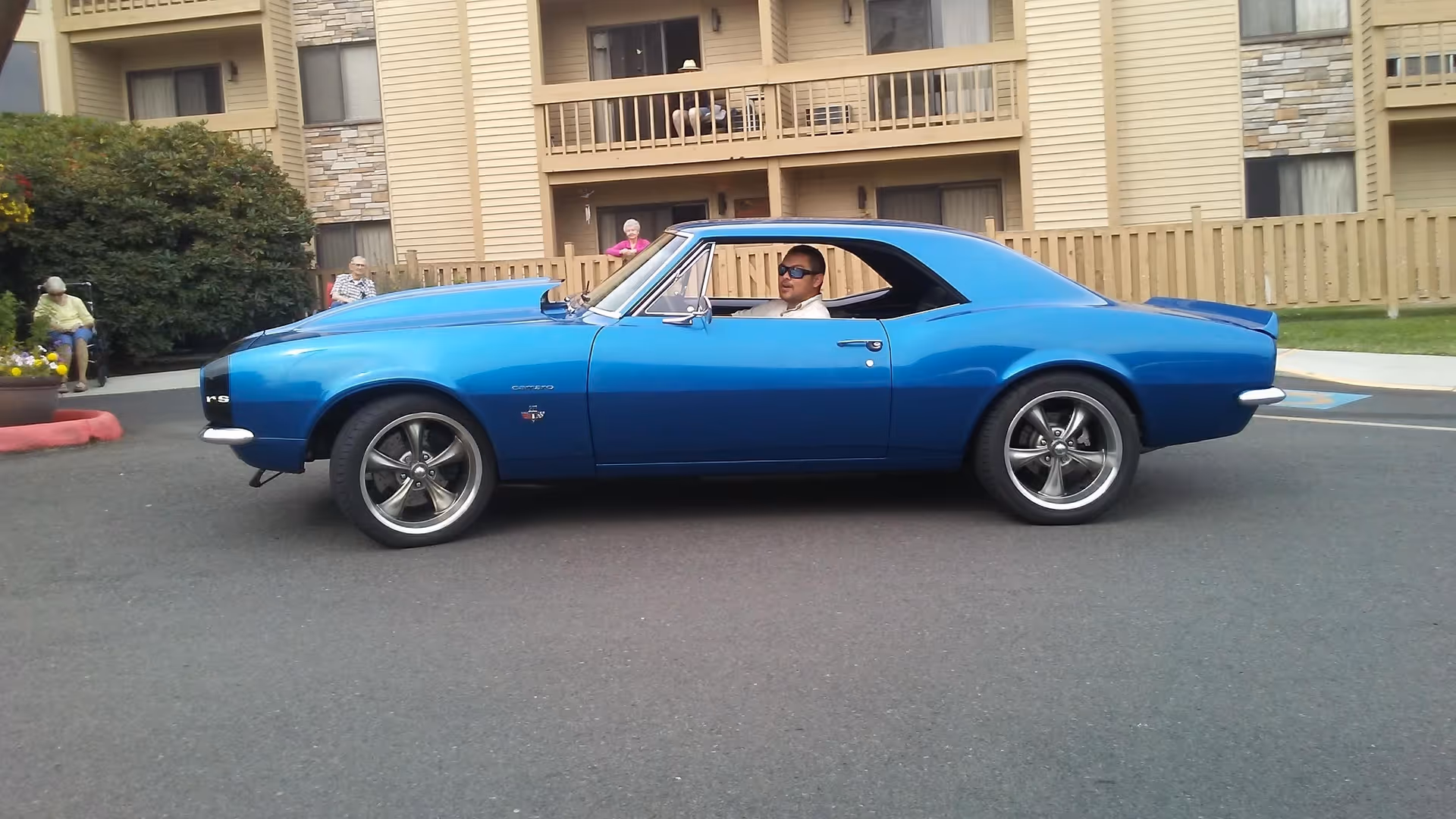 A bright blue classic car parked on a paved area in front of a beige multi-story assisted living building. A man wearing sunglasses is sitting inside the car. Several elderly people are sitting and standing near the building in the background.