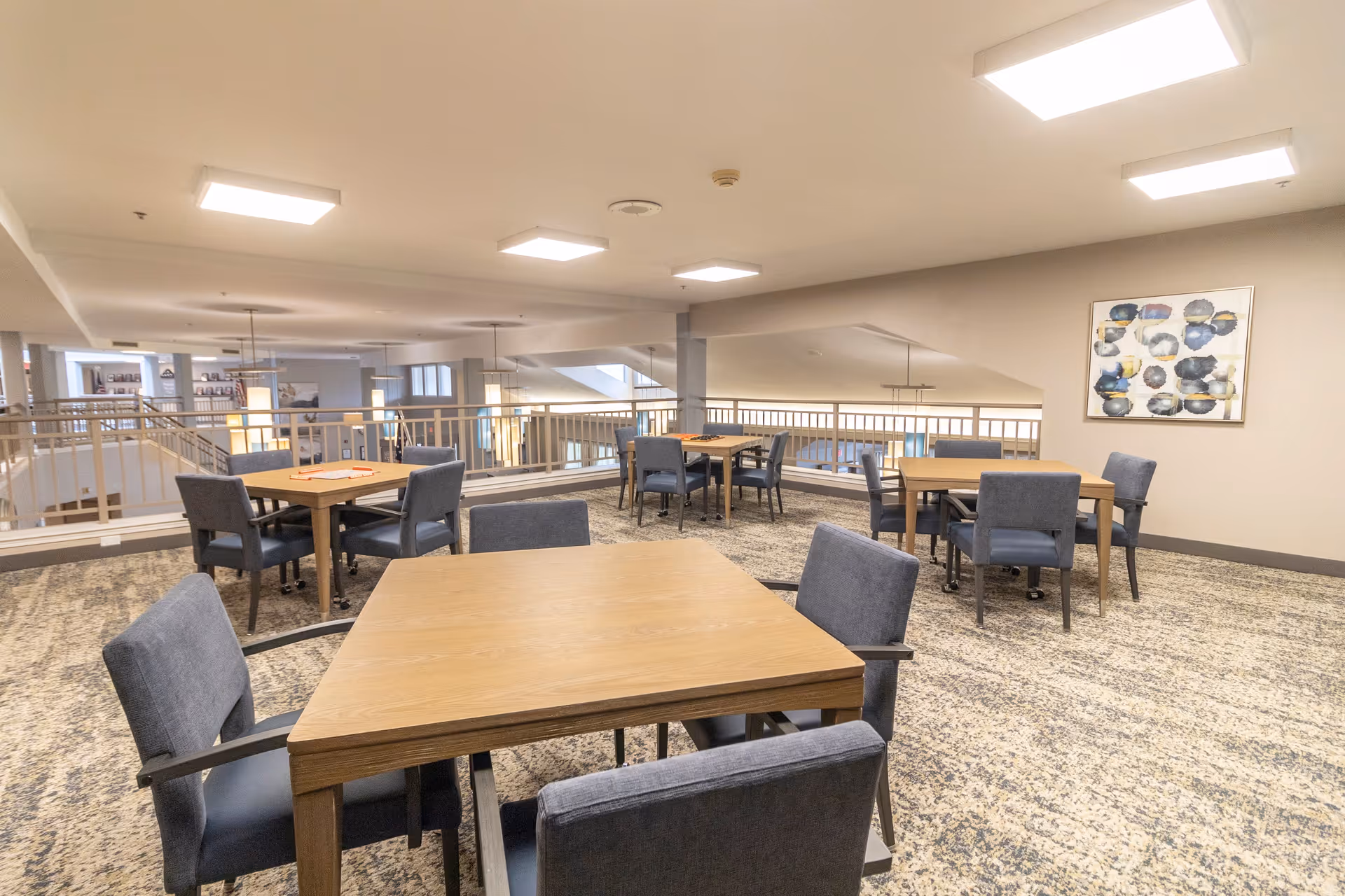 A spacious, well-lit common area with multiple wooden tables and blue upholstered chairs arranged for seating. The room has a patterned carpet, neutral-colored walls, modern ceiling lights, and a decorative abstract painting on one wall. The area overlooks a lower level with railings and additional seating visible.