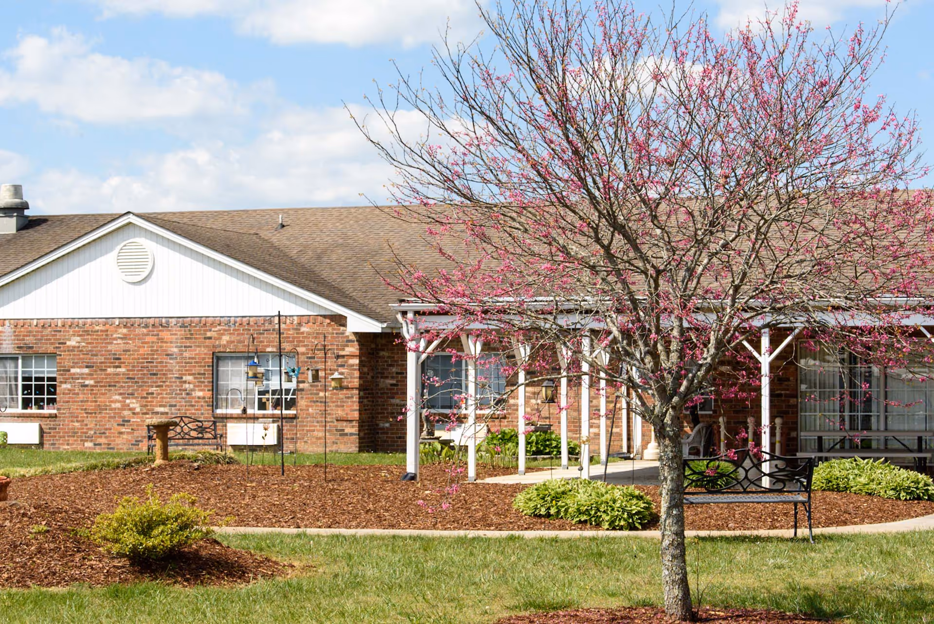 Outdoor view of a brick building with a brown roof, featuring a covered porch area with white pillars. In the foreground, there is a tree with pink blossoms, green grass, and landscaped mulch beds with small bushes. The sky is partly cloudy.