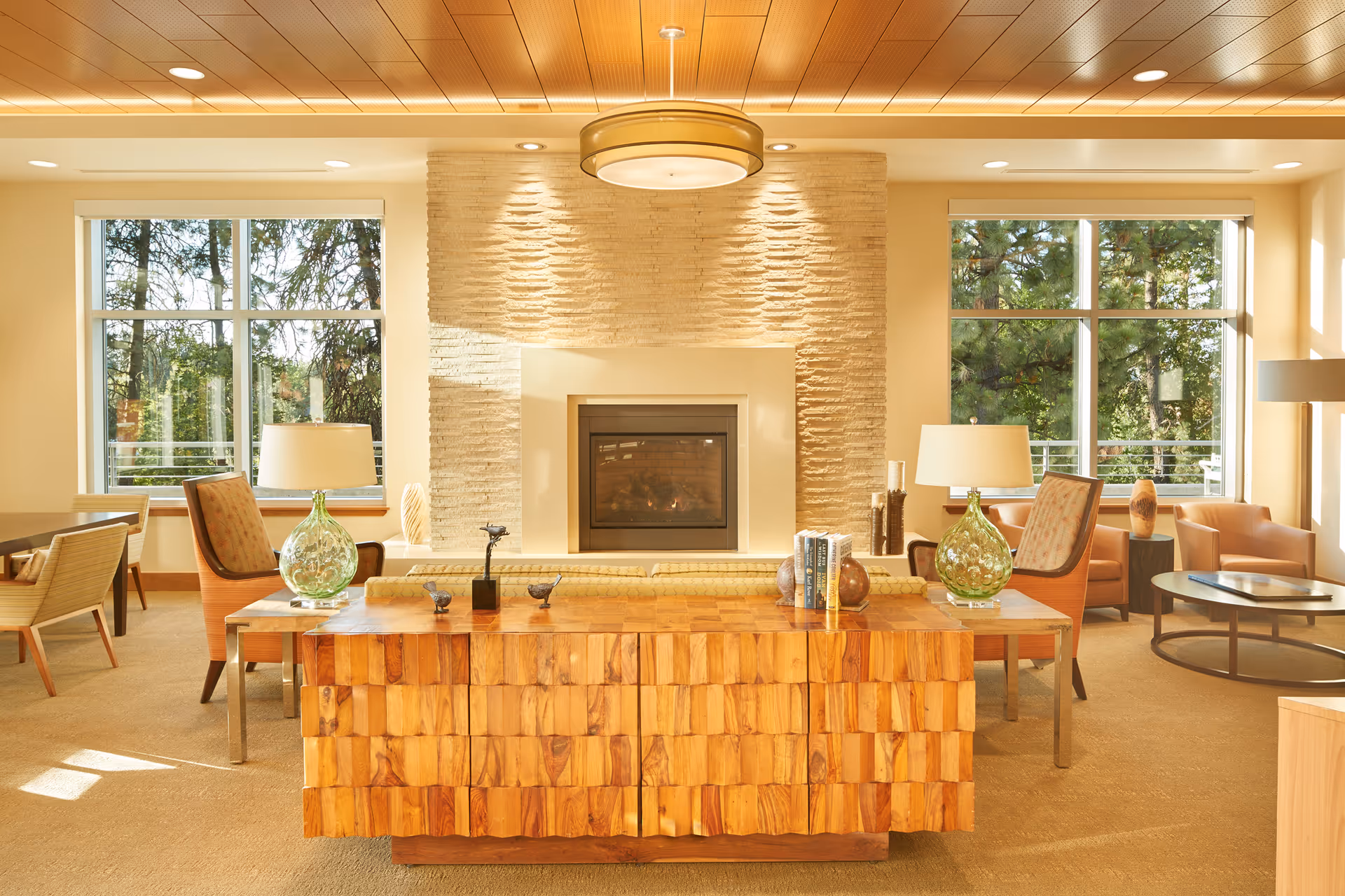 Sunlit communal living room with a central fireplace, large windows, seating areas, and a wooden console table.