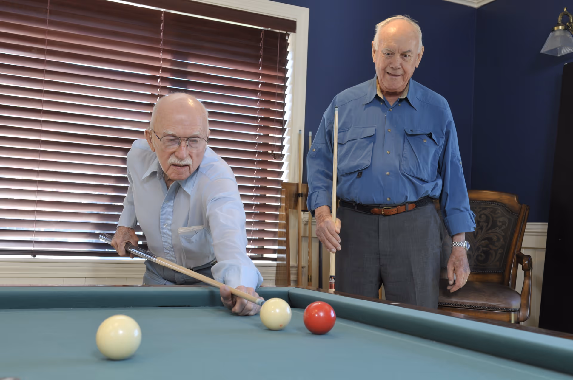 Two elderly men playing billiards indoors. One man is leaning over the pool table aiming with a cue stick, while the other stands nearby holding a cue stick and watching. The room has dark blue walls, a window with closed blinds, and a wooden chair in the background.
