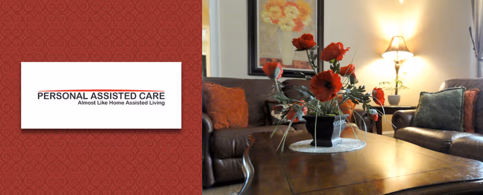 A cozy living room with brown leather sofas adorned with orange and green cushions, a wooden coffee table with a vase of red flowers, a side table with a lamp, and a framed floral painting on the wall.