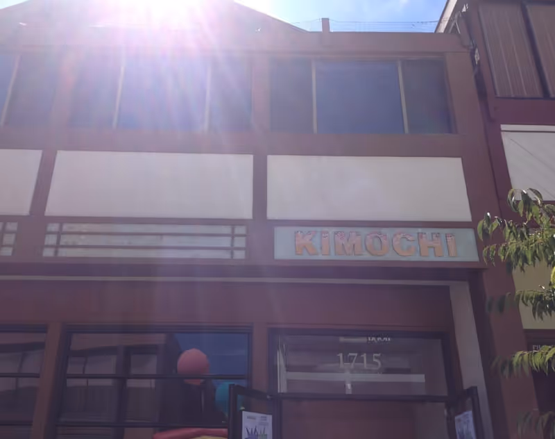 Exterior view of a building with a sign that reads 'KIMOCHI' above the entrance. The building has large windows on the upper floor and the sun is shining brightly, creating lens flare in the image.