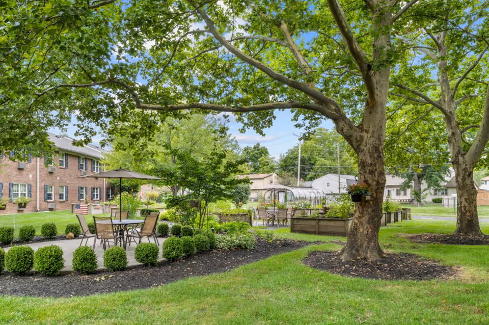 Outdoor garden area at Oakleaf Village of Columbus with green grass, large trees, trimmed bushes, raised garden beds, and patio tables with chairs and umbrellas. A brick building is visible in the background along with neighboring houses.