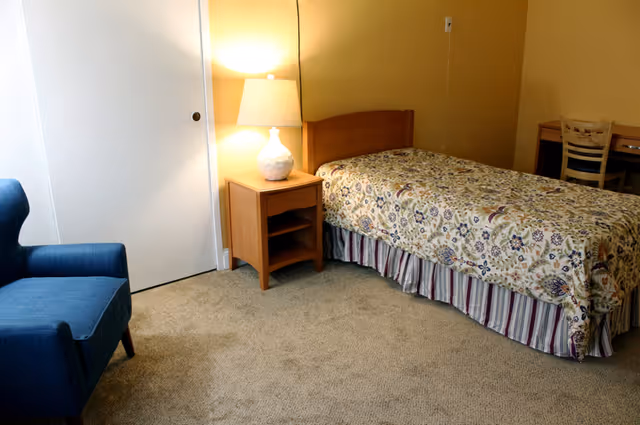 A small bedroom with a single bed covered in a floral patterned bedspread and a striped bed skirt. Next to the bed is a wooden nightstand with a white lamp on it. There is a blue upholstered armchair to the left and a wooden desk with a chair in the background. The room has beige walls and carpeted floor.