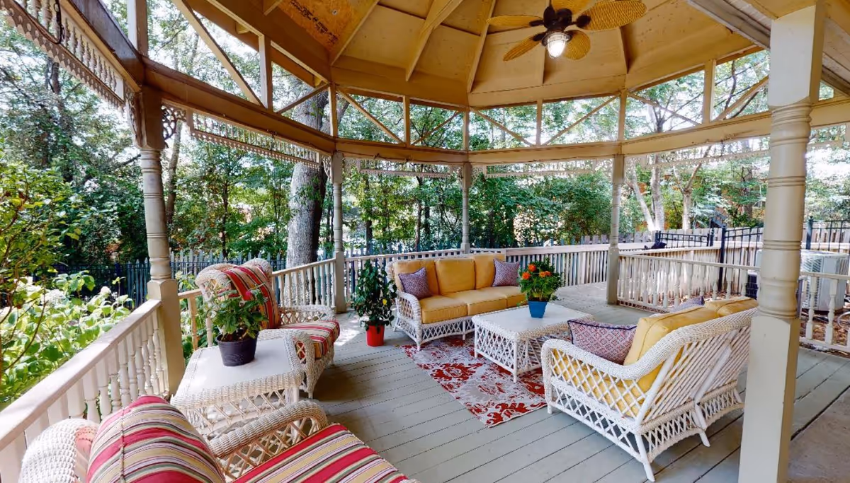 Covered gazebo-style porch with white wicker seating, colorful cushions and plants under a ceiling fan overlooking trees.